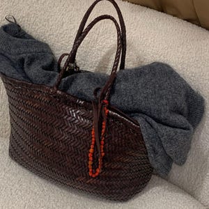 May include: A dark brown woven leather tote bag with two braided handles and a decorative orange beaded charm hanging from the front. A soft, charcoal grey knit sweater is draped inside the bag, partially covering the interior opening.