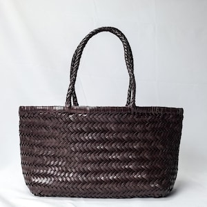 Può includere: Una borsa tote marrone scuro caratterizzata da una struttura intrecciata su tutto il corpo e due manici intrecciati. La borsa presenta una base rettangolare e un'ampia apertura superiore, con un motivo a spina di pesce in tonalità cioccolato.