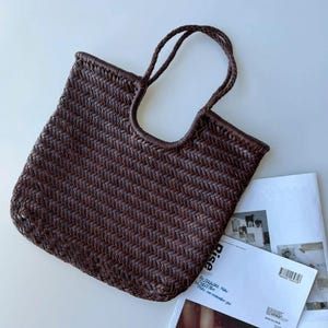 May include: A dark brown tote bag constructed from woven leather strips. The bag features a rectangular body with a flat base and two integrated braided leather handles that extend from the top opening. The weave pattern is consistent throughout.