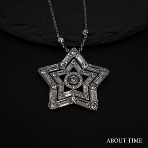 Puede incluir: Un collar con colgante de estrella plateada con una cadena fina. El colgante está adornado con cristales transparentes y brillantes de varias formas, incluyendo baguette y redondos. El collar se muestra sobre un fondo negro. El texto "ABOUT TIME" es visible en la parte inferior derecha.