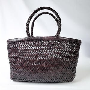 Puede incluir: Un bolso de mano tejido en color marrón oscuro con un cuerpo rectangular estructurado, un patrón de espiga y dos asas trenzadas. El bolso está construido con tiras finas entrelazadas, creando una textura uniforme en todo el exterior.