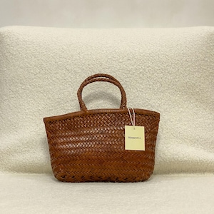Puede incluir: Un bolso de mano de cuero trenzado marrón con dos asas trenzadas y una base rectangular. Una etiqueta rectangular de color amarillo claro con el texto Margaret Lv está sujeta a la parte frontal del bolso con un cordel blanco fino.