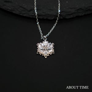 Puede incluir: Un collar plateado con un colgante de copo de nieve. El colgante está hecho de cristales transparentes y brillantes dispuestos en forma de estrella de seis puntas. La fina cadena cuelga sobre un fondo negro. El texto "ABOUT TIME" está en la parte inferior.