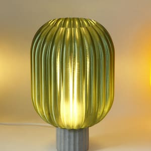 Puede incluir: Una lámpara de mesa moderna con una pantalla acanalada de color verde oliva y una base de hormigón gris. La lámpara emite un brillo cálido y acogedor. La pantalla tiene ranuras verticales y la base es cilíndrica.
