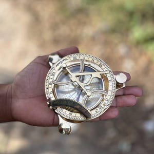 May include: A detailed brass sundial, displaying Roman numerals and a compass design. The sundial is held in a hand, highlighting its polished finish and vintage style. The text "West London" is visible on the sundial.