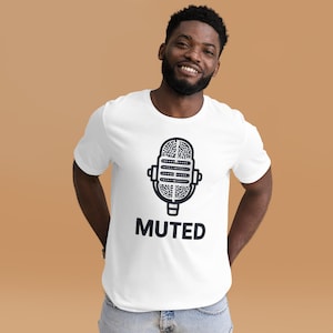 Puede incluir: Camiseta blanca con un gráfico de micrófono negro y la palabra "MUTED" debajo. El diseño del micrófono está detallado con líneas y formas. La camiseta es de cuello redondo.