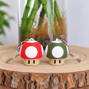 Può includere: Due portachiavi a forma di fungo, uno con cappello rosso e uno con cappello verde oliva. Entrambi presentano macchie circolari bianche sul cappello, un gambo beige e due piccoli segni rettangolari neri sulla parte anteriore con un anello.