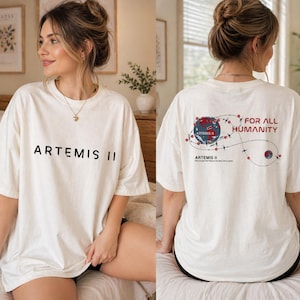 Peut inclure: T-shirt crème avec l'inscription noire "ARTEMIS II" sur le devant. Le dos présente un graphique de la Terre, une trajectoire de vol et les mots "FOR ALL HUMANITY" et "ARTEMIS II".