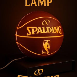 May include: A spherical basketball lamp featuring the Spalding logo and NBA emblem illuminated in orange light. The lamp sits on a dark rectangular base with a white power cord. The text BASKETBALL LAMP and the Spalding logo appear above and below.