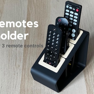 May include: A black and cream remote control holder designed for three devices. The holder features three vertical slots and includes the text remotes holder for 3 remote controls printed on the left side of the image in white and grey.