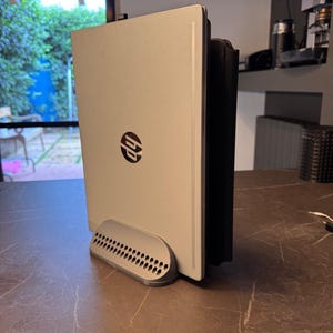 May include: A silver HP laptop standing vertically in a grey 3D-printed plastic dock. The laptop is closed, displaying the circular HP logo in the center of the lid. The dock features a curved front edge with a grid of small circular ventilation holes.