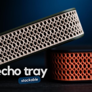 May include: Two stackable trays with a dark frame and a light-colored, intricate, repeating lattice pattern on the side. The top tray is olive green with a beige lattice, and the bottom tray is black with an orange lattice. Text reads echo tray stackable.