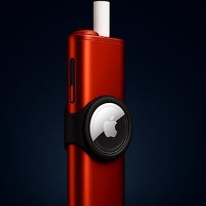 May include: A metallic red rectangular device with a white cylindrical top protrusion and a black circular holder attached to the side. The holder contains a silver Apple AirTag featuring the iconic apple logo and circular text on its surface.