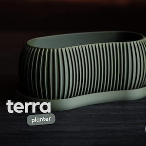 May include: An oval-shaped sage green planter with a ribbed exterior texture and a matching base tray. The text "terra planter" is printed in white on the left, and a circular green logo with the text "h3liø design" is in the bottom right corner.