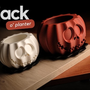 May include: Two pumpkin-shaped planters, one white and one orange, featuring carved facial expressions with large circular eyes and stitched mouths. The text "jack o' planter" appears in the upper left corner, and a circular logo is in the bottom right.