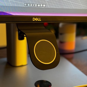May include: A black magnetic mount attached to the underside of a Dell computer monitor. The mount features a circular face with a gold-colored ring accent. The monitor stand base is visible below, and the screen displays a digital grid interface.