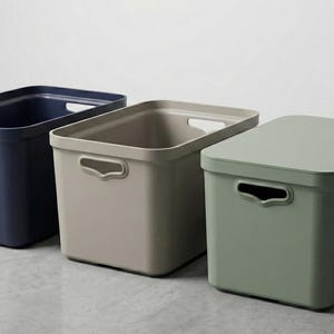 May include: Three rectangular storage bins with integrated handles on the sides. The first bin is dark navy blue, the second is a neutral beige, and the third is sage green with a matching lid placed securely on top of the container.