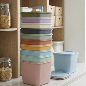 May include: A vertical stack of ten square storage containers with lids in various muted colors including olive, lavender, cream, charcoal, sage, terracotta, mustard, sky blue, mint, and dusty rose, with one additional sky blue container nearby.