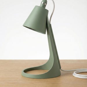 May include: A sage green desk lamp with a conical shade and a curved base, featuring a white power cord. The lamp includes text at the bottom that reads: PREDISPOSED FOR E14 BULB HOLDER - Modern Design, Versatile Use. The lamp has a matte finish.