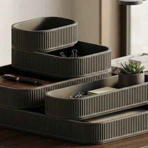 May include: A set of dark olive green desk organizers with a vertical ribbed texture. The modular system includes multiple rectangular trays of varying heights and sizes, stacked to hold office supplies like a pen, paper, and a small succulent plant.