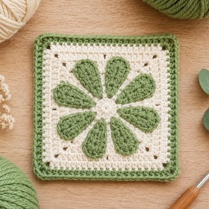 Op de afbeelding: Een vierkant haakmotief met een centraal bloemdesign met acht saliegroene bloemblaadjes op een crèmekleurige achtergrond, omlijst door een saliegroene rand. Het stuk is gemaakt met zichtbare garensteken en toont een geometrisch bloempatroon.