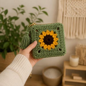 May include: A square crochet coin purse featuring a sunflower motif with a dark brown center and bright yellow petals, surrounded by sage green yarn. The pouch includes a gold-toned metal zipper closure and a matching sage green yarn tassel.