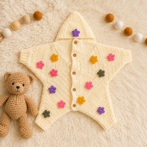 May include: A cream-colored, star-shaped hooded garment featuring a front button closure and small, multi-colored crocheted flower appliqués in shades of pink, yellow, purple, and green, accompanied by a small, light brown crocheted teddy bear.