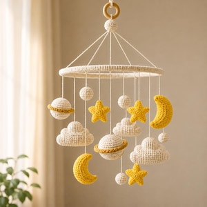 May include: A hanging mobile featuring crocheted elements in cream and yellow. The design includes two planets with rings, two crescent moons, three stars, three clouds, and three small spheres suspended from a circular frame with a wooden ring.