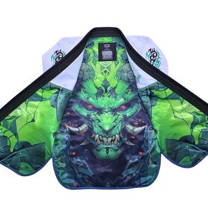 Puede incluir: Gi de artes marciales blanco y negro con un vibrante gráfico de monstruo verde y azul en el interior. El gi tiene ribetes negros y diseños tribales en los hombros. El interior presenta una ilustración detallada de un monstruo.
