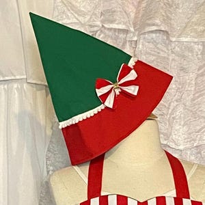 May include: A green and red elf hat with a white trim and a bow. The hat is on a mannequin. The hat is part of a Christmas costume.