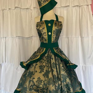 May include: A green and gold dress with a matching hat. The dress features a halter top, a fitted bodice with gold buttons, and a full skirt with gold trim. The hat has a bow and gold trim. The fabric has a floral pattern.
