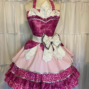 May include: A pink and white apron with a large bow and a ruffled skirt. The apron has a white and silver swirl pattern and a pink and white polka dot pattern.