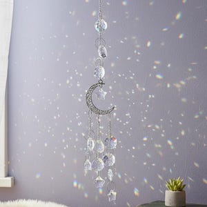 Puede incluir: Un atrapasoles plateado con forma de luna y estrella cuelga de una cadena, adornado con cuentas y colgantes de cristal transparente. La decoración de temática celestial está suspendida frente a una pared de color púrpura claro, con reflejos de luz que crean un efecto brillante.
