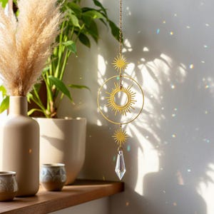May include: A gold-toned suncatcher with a celestial design. It features a sunburst within a circle, star accents, and a clear crystal prism. The suncatcher hangs from a chain, creating a decorative effect.