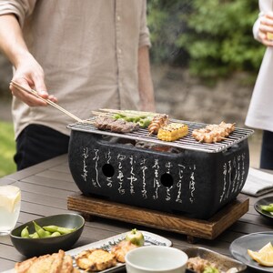 Può includere: Un barbecue rettangolare in ceramica nera con calligrafia giapponese bianca e fori di ventilazione circolari. Il barbecue poggia su una base in legno ed è sormontato da una griglia metallica con spiedini di carne, mais e verdure verdi.