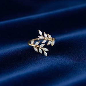 May include: A gold-toned open ring featuring two overlapping branches adorned with marquise-cut clear stones. The design mimics the appearance of leaves, with the stones set along the delicate metal stems that curve to form the band of the ring.