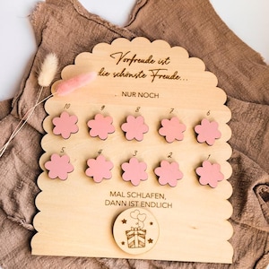 Puede incluir: Tablero de cuenta regresiva de madera con borde festoneado, con marcadores rosas en forma de flor numerados del 1 al 10. El tablero tiene texto grabado en alemán. Una pequeña pieza redonda de madera con una caja de regalo y globos está en la parte inferior.