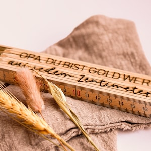 May include: A wooden folding ruler with the text "OPA DU BIST GOLD WERT au jedem Zentimeter" is displayed. The ruler is partially open, showing centimetre markings. Dried wheat and a tan cloth are in the background.