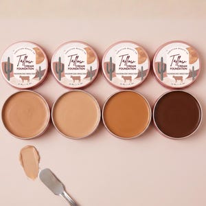 May include: Four round metal tins of Christian Beauty Co. Tallow Cream Foundation in varying shades, each with a label featuring a cactus, sun, and cow graphic. A metal spatula rests nearby with a smear of light beige foundation cream on the surface.