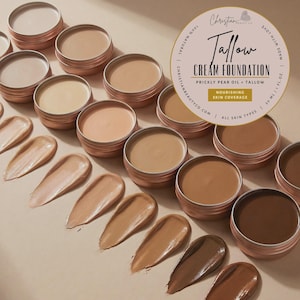 Könnte beinhalten: Eine Auswahl an Tallow Cream Foundation in verschiedenen Farbtönen, präsentiert in kleinen, runden, kupferfarbenen Dosen. Unter den Dosen werden Farbmuster der Foundation angezeigt. Das Produkt ist mit dem Text "Tallow Cream Foundation" und "Prickly Pear Oil + Tallow" beschriftet.