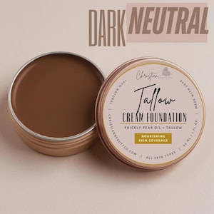 May include: An open, round, metal tin of dark neutral-toned cream foundation. The label reads "Tallow Cream Foundation" with "Prickly Pear Oil + Tallow" and "Nourishing Skin Coverage". The text "Dark Neutral" is at the top.