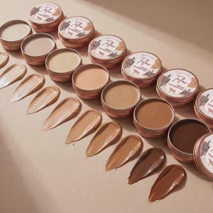 May include: A collection of round copper-coloured metal tins containing cream foundation in various shades, ranging from light beige to deep brown. Each lid features a label with the text Tallow Cream Foundation and a cactus and cow graphic.