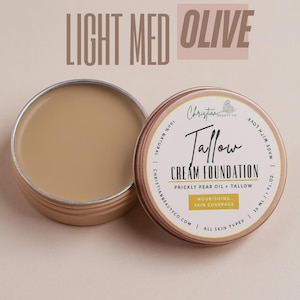 May include: An open, round, light brown tin of cream foundation next to a closed tin. The closed tin has a label with the text "Tallow Cream Foundation" and "Nourishing Skin Coverage". The text "Light Med Olive" is at the top.