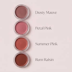 Puede incluir: Cuatro recipientes circulares de producto cosmético en crema dispuestos verticalmente. Los tonos están etiquetados como Dusty Mauve, Petal Pink, Summer Pink y Rum Raisin. Cada recipiente tiene una tapa transparente y una superficie lisa.