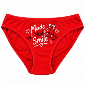 以下が含まれることがあります： 鮮やかな赤色の下着で、前面中央に白い文字で「Made You Smile」とプリントされ、白いリボンが付いた2つのサクランボのグラフィックと、いくつかの小さな白いハートの輪郭が描かれた、クラシックなブリーフカットのデザインです。
