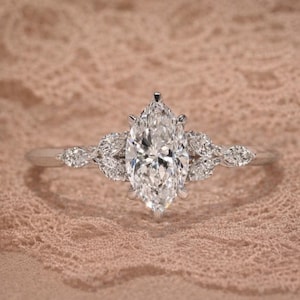 Op de afbeelding: Een verlovingsring met een marquise-geslepen diamant en een delicate band. De ring heeft een grote, sprankelende centrale steen en kleinere accentdiamanten. De zetting is helder, zilverkleurig gepolijst.