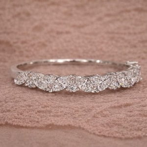 Può includere: Un anello d'argento con una fila di pietre preziose taglio marquise. Le pietre sono incastonate vicine, creando una fascia continua di brillantezza. L'anello è posto su uno sfondo rosa-marrone chiaro.