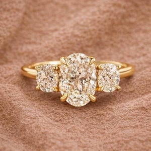 May include: A gold ring featuring three large, sparkling diamonds. The center diamond is oval-shaped, flanked by two cushion-cut diamonds. The ring is set on a simple gold band, showcasing the brilliance of the stones.