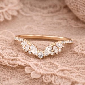 Puede incluir: Un anillo dorado con un diseño curvo. El anillo presenta una fila de pequeñas piedras transparentes a lo largo de la banda y un grupo central de piedras más grandes, transparentes, con corte marquesa. Se engastan piedras pequeñas y transparentes adicionales alrededor de las piedras más grandes.