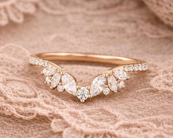 May include: A gold-colored ring with a curved design. The ring features a row of small, clear stones along the band and a central cluster of larger, clear, marquise-cut stones. Additional small, clear stones are set around the larger stones.